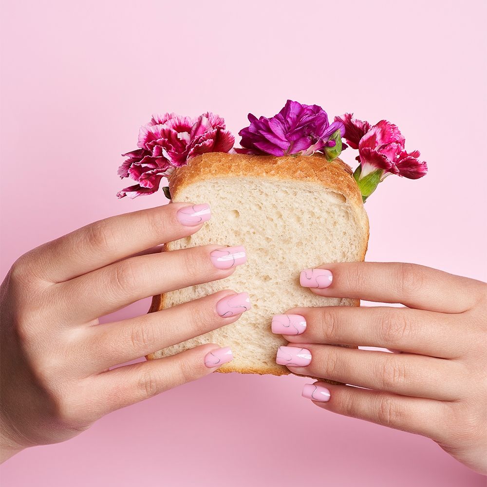 Hände mit pinkem Nageldesign halten ein Stück Brot, aus dem Blumen wachsen
